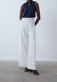 Granatowa bezrękawnik z dekoltem typu keyhole z przodu, w zestawieniu z białymi spodniami o wysokim stanie i szerokich nogawkach oraz czarnymi sandałami na obcasie.