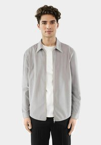 Lichtgrijze ritsjack van gladde stof, met een puntige kraag en lange mouwen, gedragen over een wit shirt. Zwarte broek maakt de look compleet.