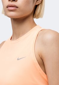 Κοντινό πλάνο ενός ατόμου που φοράει ένα αμάνικο ροδακινί Nike tank top με ένα μικρό ασημένιο σήμα Nike στο στήθος.