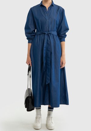 Mujer con un vestido largo de denim azul con un cinturón atado en la cintura, zapatillas blancas, calcetines a rayas y sosteniendo un bolso negro con una correa de cadena.
