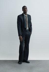 Completo nero con collo sciallato a contrasto, cravatta a fantasia e camicia a quadri. Il tessuto sembra liscio; i pantaloni sono sartoriali e aderenti.