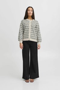 Gilet en maille crème avec motif de nœud noir, doté d'un devant à boutons et de manches ballons, associé à un pantalon large noir et des sandales.
