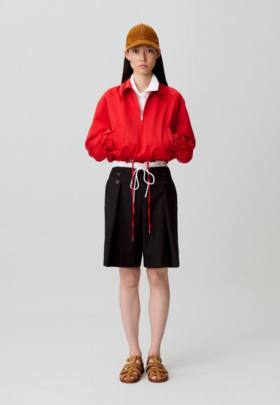 Giacca rossa con cerniera e polsini elasticizzati, pantaloncini neri con dettagli a bottoni e sandali marroni con cinturini, abbinati a un berretto marrone.