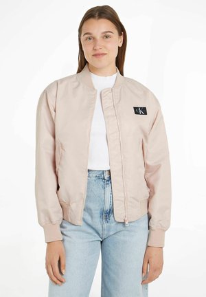 Giacca bomber rosa chiaro con zip frontale, polsini a costine e una patch nera sul petto. Indossata sopra una maglietta bianca e jeans in denim chiaro.