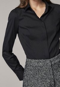 Camicia nera a maniche lunghe con bottoni, colletto e design aderente, abbinata a una gonna grigia con texture e motivo.
