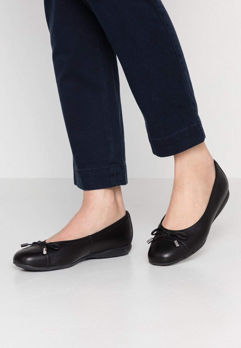 Bailarinas de cuero negro con punta redondeada, detalle de lazo en la parte delantera y un acabado suave. Combinadas con pantalones azul marino.
