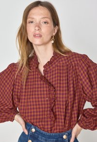 Blusa a quadri rossi e marroni con scollatura a rouches e bottoni, abbinata a una gonna in denim blu con bottoni dorati.