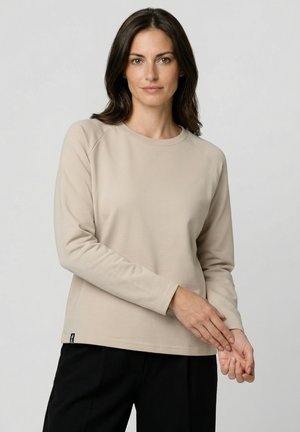 Femme aux cheveux foncés portant un haut beige à manches longues et un pantalon noir, debout devant un fond gris clair uni.