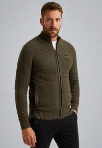 Cardigan zip-up a maglia di colore verde oliva scuro, con motivo testurizzato, colletto alto e due tasche laterali. Dettaglio a toppa sul petto.