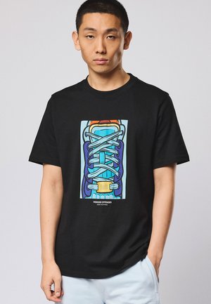 Jonge man die een zwart T-shirt draagt met een kleurrijke veterprint en de tekst "WEEKEND OFFENDER ADMIT NOTHING" op de voorkant.