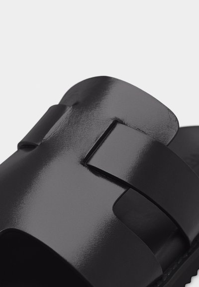 Κοντινό πλάνο μαύρου γυαλιστερού σανδαλιού τύπου slide με φαρδιά λουρί που διαθέτει ορθογώνια λεπτομέρεια κοπής.