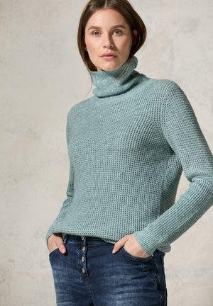 Pullover in teal chiaro lavorato a maglia con collo alto, motivo testurizzato e maniche lunghe, abbinato a jeans in denim scuro con dettagli a bottoni.