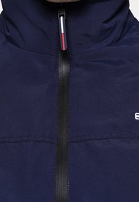 Tmavě modrá bunda s vysokým límcem, černým zipem a malým bílým logem. Má hladkou texturu a viditelné švy.