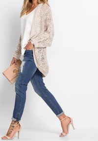 Cardigan aperto beige su una maglietta bianca, abbinato a jeans blu strappati e sandali con tacco nudo. Gli accessori includono una pochette color beige chiaro.