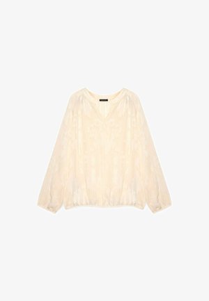 Blusa in crema con maniche lunghe, scollo a V, orlo elastico e un sottile motivo floreale su tessuto trasparente. La finitura texture aumenta l'interesse visivo.