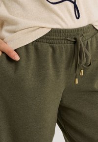 Pantalon de survêtement vert olive avec taille élastique et cordon de serrage, avec embouts métalliques dorés. Texture douce, design décontracté, avec poche latérale visible.