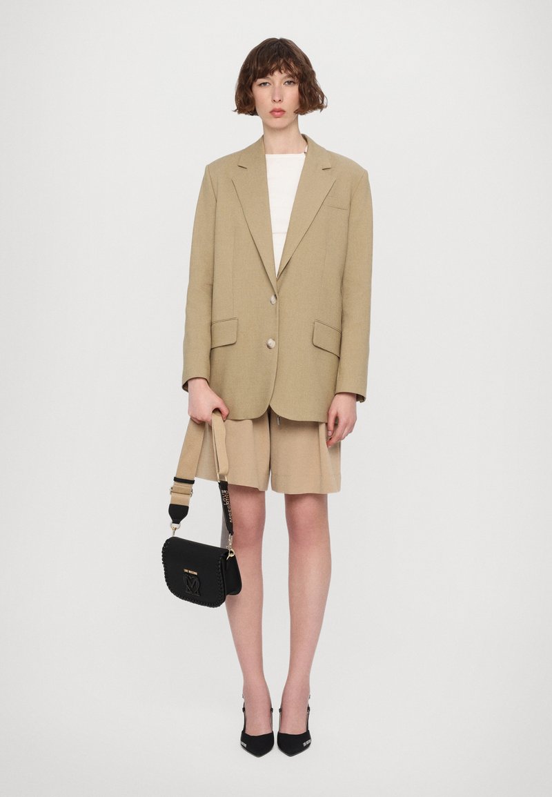 Blazer oversized beige con due bottoni, abbinato a pantaloni corti beige. La modella tiene una borsa a tracolla nera con dettagli testurizzati e indossa tacchi neri.