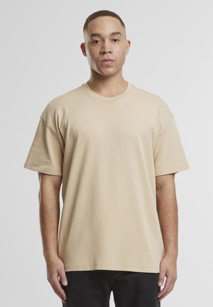 Homme aux cheveux courts portant un t-shirt beige oversize et un pantalon noir, se tenant devant un fond uni gris clair.