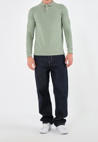 Lange mouw lichtgroen poloshirt met een knoopsluiting kraag, gecombineerd met donkerblauwe jeans en witte sneakers, tegen een effen achtergrond.