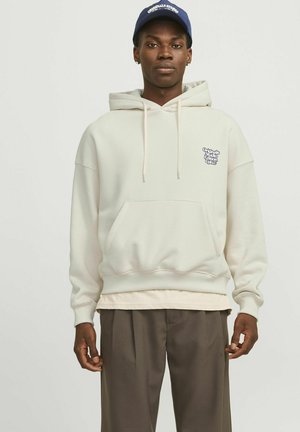 Hoodie in off-white katoenmix, met een voorzak en een klein logo op de borst. Ggedragen over een lichtkleurig shirt.