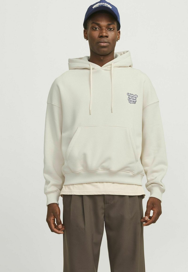Sudadera con capucha de mezcla de algodón en color blanco roto, que cuenta con un bolsillo frontal y un pequeño logotipo en el pecho. Lleva puesta una camiseta de color claro debajo.