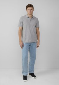 Polo gris avec un col, des manches courtes et trois boutons, associé à un jean bleu clair et des baskets noires, se tenant devant un fond neutre.