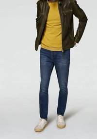 Donkergroene leren jas, mosterdkleurige trui, blauwe slim-fit jeans, beige sneakers met witte veters, staande tegen een neutrale achtergrond.
