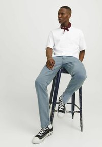 Un tricou alb cu mânecă scurtă, blugi albaștri deschiși, adidași negri cu talpă albă și o batică burgundă în jurul gâtului, așezat pe un taburet.