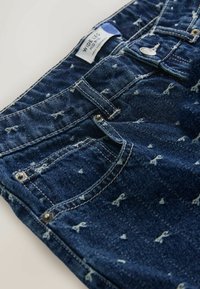 Plačiąsias džinsines kelnes tamsiai mėlynos spalvos su subtiliu šviesiai mėlynu siuvinėjimo raštu. Yra kišenės, metalinė sagčia ir siuvinėtos siūlės.