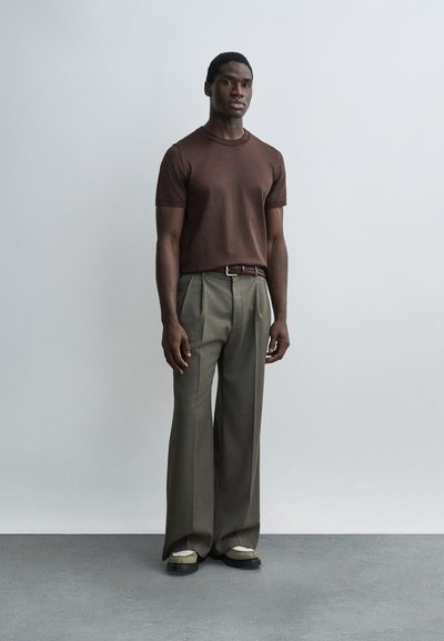 Άνδρας που στέκεται μπροστά σε απλή γκρι ανοιχτή τοίχο, φοράει καφέ κοντομάνικο πουκάμισο, χακί παντελόνι με φαρδιά μπατζάκια και δίχρωμα παπούτσια.