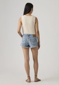 Γυναίκα με μακριά σκούρα μαλλιά, που φοράει αμάνικο μπεζ τοπ, μπλε τζιν σορτς με ξεφτισμένα άκρα και καφέ δερμάτινα σανδάλια με κορδόνια, στέκεται με την πλάτη στραμμένη.