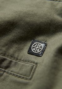 Olivgrön tyg med en strukturerad yta, med en svart rektangulär patch där bokstäverna "JCKS" är präglade i silver och en liten ficka.