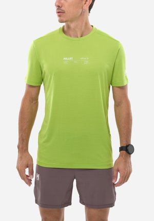 Homme portant un t-shirt à manches courtes Millet vert citron et un short marron, regardant vers la gauche, avec une montre connectée noire au poignet gauche et une boucle d'oreille à l'oreille droite.