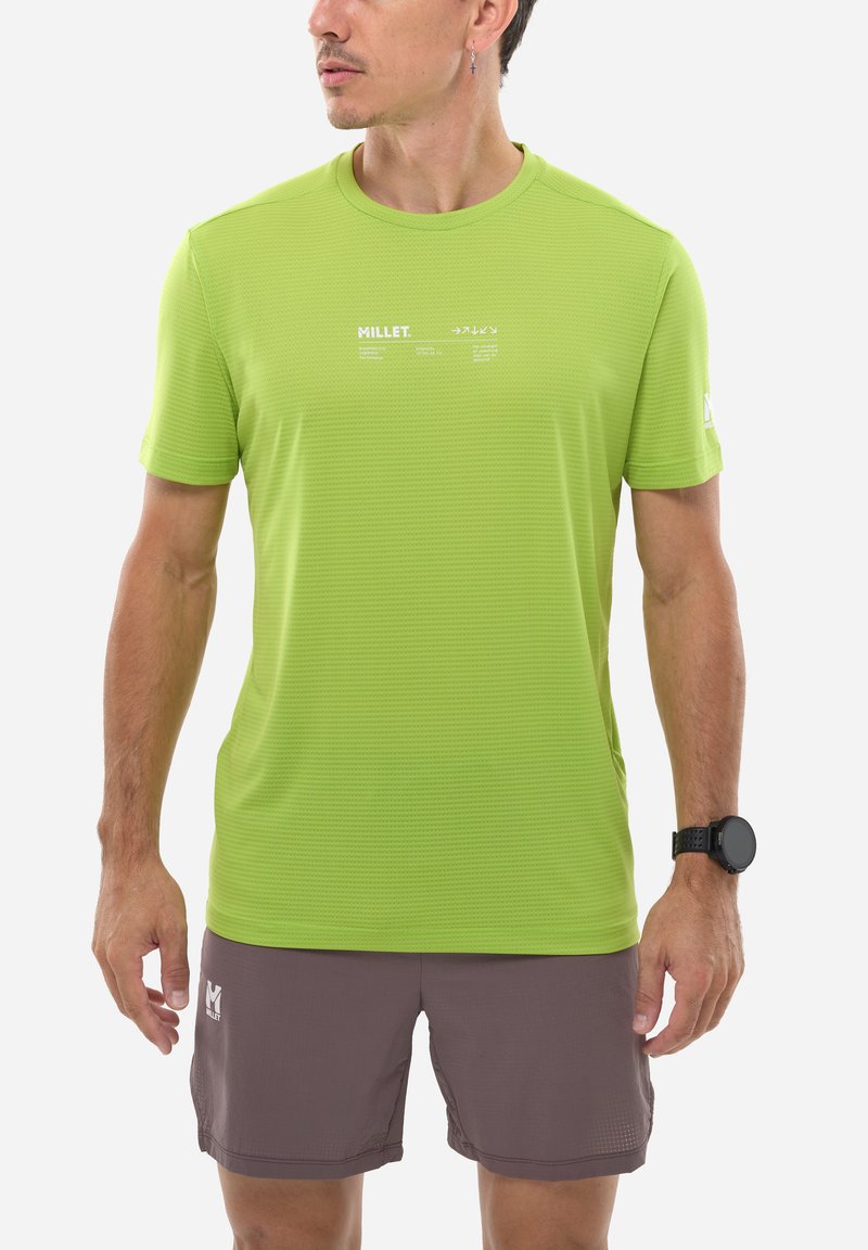Homme portant un t-shirt à manches courtes Millet vert citron et un short marron, regardant vers la gauche, avec une montre connectée noire au poignet gauche et une boucle d'oreille à l'oreille droite.