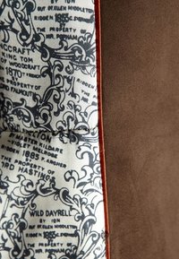 Material de ante marrón por un lado, contrastando con una tela estampada que presenta texto azul y diseños ornamentados por el otro, separados por un delgado ribete naranja.