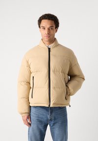 Jachetă puffer bej cu guler înalt, fermoar frontal și fermoare negre în contrast. Dispune de mansete elastice și un design cusut.