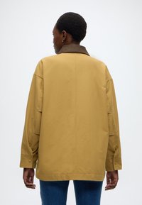 Veste oversize jaune moutarde avec un col marron, des poignets à boutons et une texture lisse, présentant une coupe droite et sans motifs visibles.