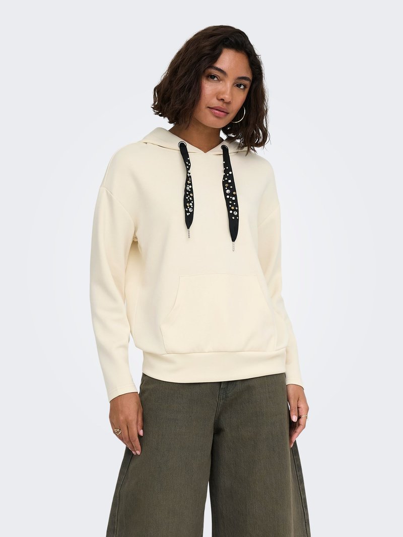 Femme aux cheveux foncés mi-longs portant un sweat à capuche crème avec des cordons décoratifs noirs et un pantalon large vert olive.