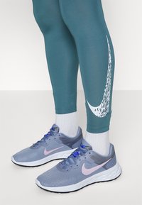 Türkise Leggings mit weißem Nike-Logo, kombiniert mit grauen Sneakers mit rosa Akzenten und strukturiertem Mesh-Obermaterial. Weiße Socken sind sichtbar.