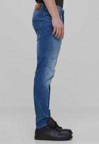 Jeans de mezclilla azul con textura desgastada, ajuste slim, cintura de tiro medio y parche de cuero marrón en la cintura trasera, combinados con zapatillas negras.