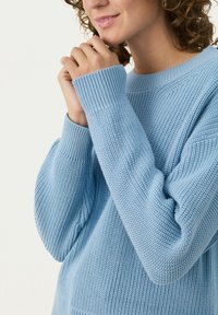 Pull en maille bleu clair avec des poignets côtelés et un motif texturé. Caractéristiques d'un col rond et d'une coupe ample autour des épaules et des bras.