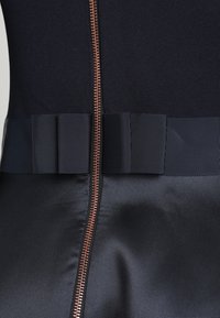 Gros plan sur un tissu foncé avec une fermeture éclair en cuivre centrale et un ruban côtelé horizontal près de la couture.