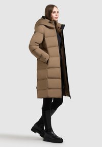 Brązowa kurtka puchowa z kapturem, z pikowanym wzorem, stojącym kołnierzem i zamkiem z przodu. Nosi ją na czarnych legginsach i kozakach za kolano.