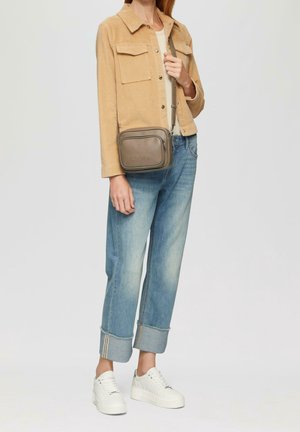 Person iført beige jakke, optrukne blå jeans, hvide sneakers og bærende en taupe crossbody-taske med synlige lynlåsdetailler.