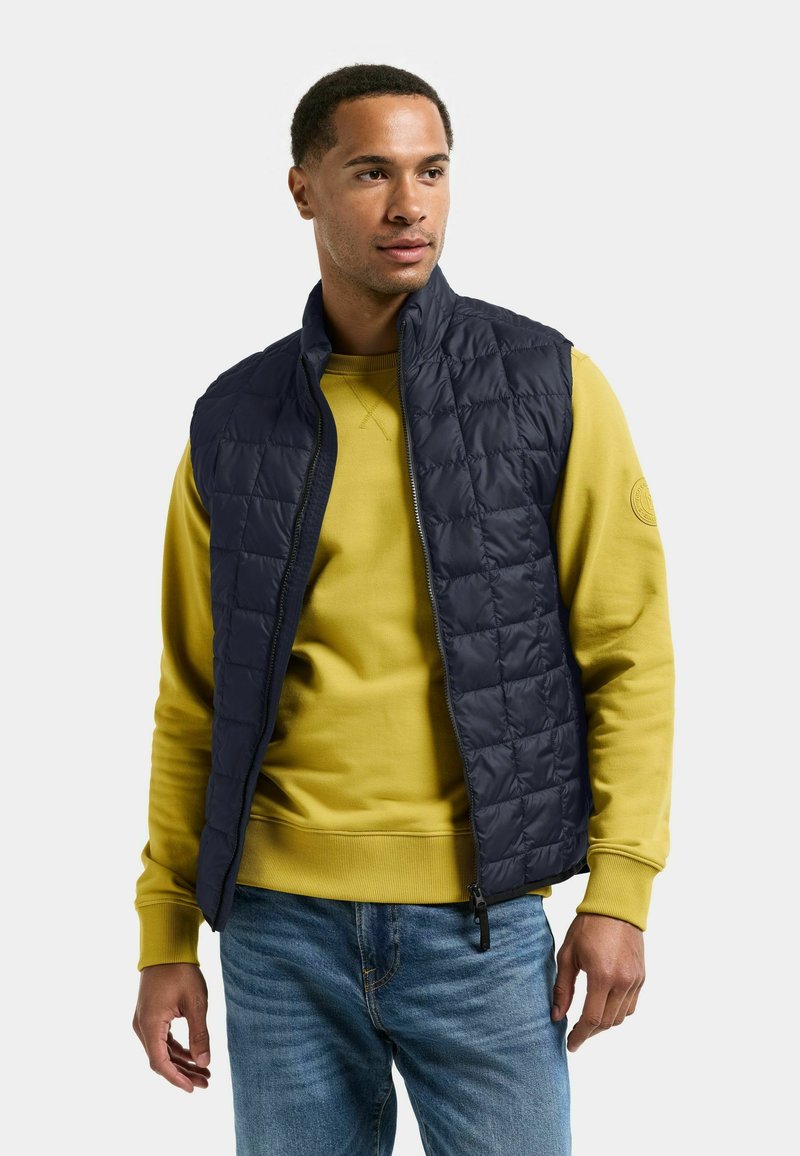 Man draagt marineblauw doorgestikt vest over mosterdkleurige trui en blauwe spijkerbroek, staat tegen een effen lichtgrijze achtergrond, kijkt naar rechts.