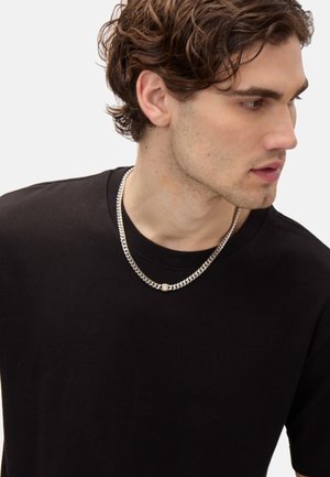Silver chain necklace with round accent, worn over a black cotton t-shirt. The chain features a flat, twisted design for texture.