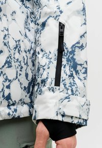 Manche de veste à motifs blancs et bleus avec une poche zippée noire. Tissu à texture lisse avec un détail de poignet. Manches longues noires visibles en dessous.