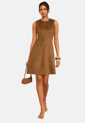 Femme portant une robe en daim marron sans manches, tenant un petit sac assorti, avec des bracelets empilés et des sandales ouvertes beiges.