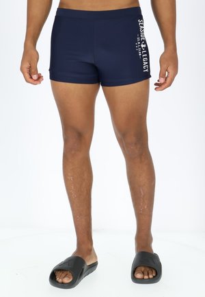 Navy swim shorts with a fitted design, made from a stretchy material. Features branding in white text on the left side. Black slide sandals.