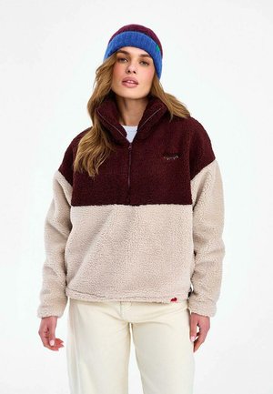 Kobieta w kremowo-bordowej polarowej kurtce z półzamkiem i niebiesko-fioletową dzianinową opaską na głowę, stoi na tle jednolitego białego tła.